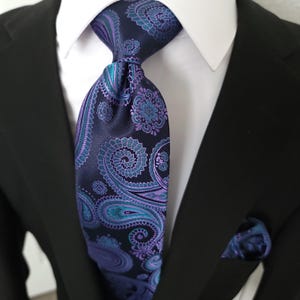 Puede incluir: Una corbata de cachemira azul marino con detalles turquesa y morado, combinada con un pañuelo de bolsillo a juego. La corbata está anudada y se usa con una camisa blanca y una chaqueta de traje negra. El estampado de cachemira presenta diseños florales y de remolino.
