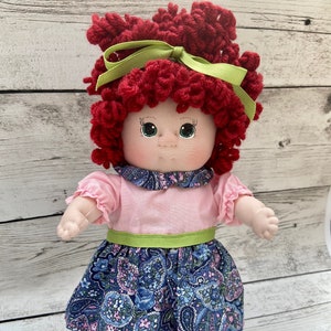 May include: A handmade doll with red curly hair, green eyes, and a pink and blue floral dress. The doll is wearing a green ribbon in her hair and white shoes.
