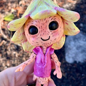 May include: A whimsical figurine of a flower fairy. The fairy has a yellow and green flower hat, a pink dress, and a smiling face with large black eyes. It stands on a green base. The figurine is hand-painted.