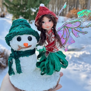 May include: A whimsical winter scene featuring a snowman and a fairy figurine. The snowman has a green crocheted hat and scarf, with a carrot nose. The fairy wears a red and green outfit with iridescent wings. The base is a wooden disc.