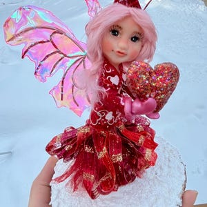 Könnte beinhalten: Handgefertigte Feenfigur mit rosa Haaren, schillernden Flügeln und einem roten Kleid. Die Fee hält ein glitzerndes Herz und hat herzförmige Akzente auf dem Kopf. Die Figur ist auf einem Holzsockel mit weißem Material befestigt.
