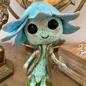 May include: A whimsical figurine of a flower fairy with a light blue flower-shaped head and large black eyes. The fairy wears a mint green dress and a matching vest with gold accents, standing on a green base. The figurine is a unique decorative item.