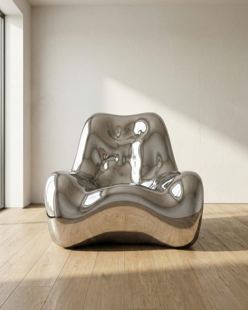 May include: A modern, sculptural chair with a reflective, metallic silver finish. The chair has a unique, flowing design with rounded edges and a low profile. It sits on a wooden floor in a well-lit room.