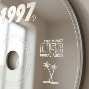 May include: A silver compact disc with the year "1997" printed in white. The disc also features the "Compact Disc Digital Audio" logo and a palm tree graphic. The disc has a reflective surface.