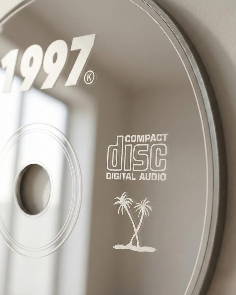 May include: A silver compact disc with the year "1997" printed in white. The disc also features the "Compact Disc Digital Audio" logo and a palm tree graphic. The disc has a reflective surface.