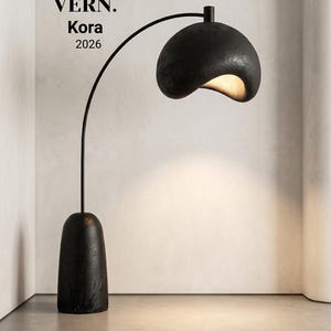 May include: A black floor lamp with a curved arm and a rounded shade. The lamp has a textured finish and a warm light emanating from the shade. The base is a dark, elongated shape. Text on the image reads "VERN. Kora 2026."