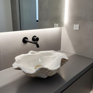 Puede incluir: Un lavabo blanco en forma de concha con un exterior beige texturizado. El lavabo se encuentra sobre una encimera gris oscuro, con un grifo negro y un espejo en el fondo. El lavabo tiene un desagüe dorado.