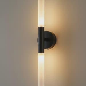 May include: A modern wall sconce with a black metal base and two vertical, frosted glass light tubes. The sconce emits a warm, inviting glow, perfect for ambient lighting in a contemporary space. The design is minimalist and sleek.
