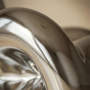 May include: Close-up of a shiny, silver metal object with a curved, flowing design. The surface reflects light, creating bright highlights and shadows. The object appears to be part of a larger piece, possibly furniture or decor.