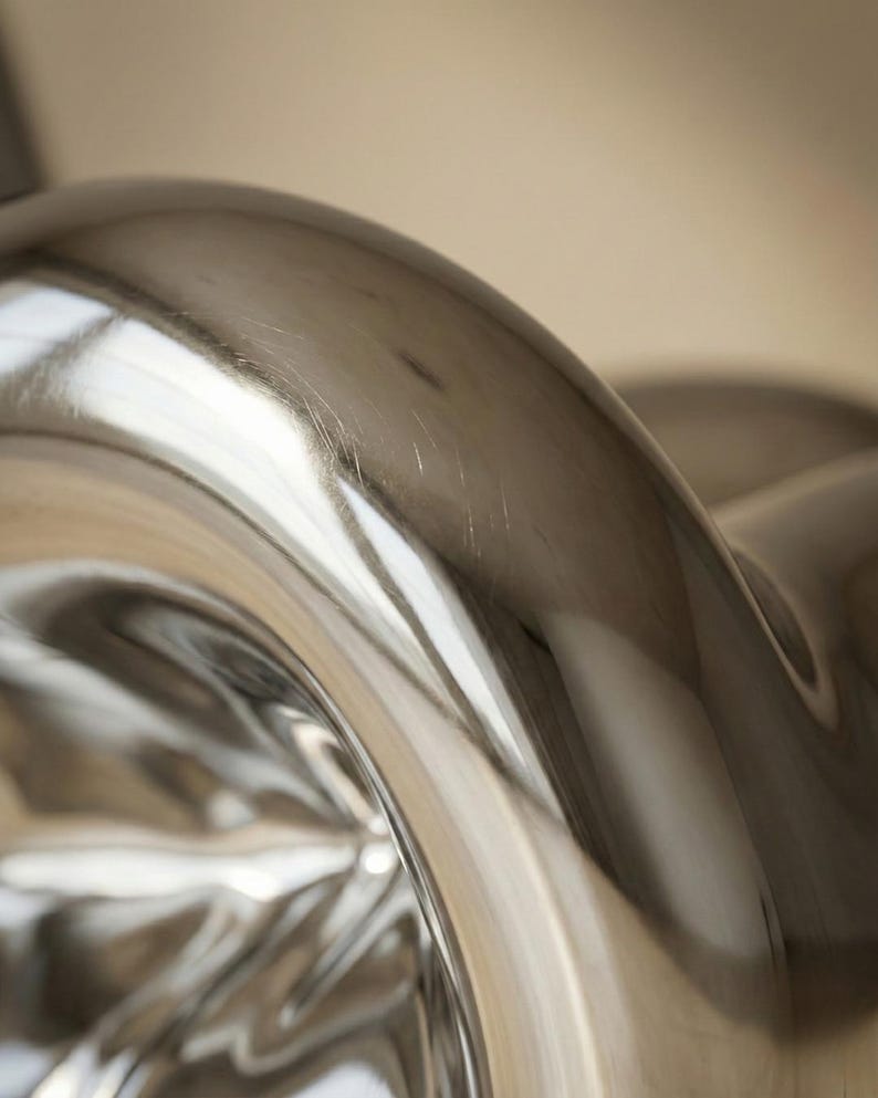 May include: Close-up of a shiny, silver metal object with a curved, flowing design. The surface reflects light, creating bright highlights and shadows. The object appears to be part of a larger piece, possibly furniture or decor.
