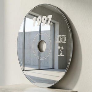May include: A reflective CD-shaped wall mirror with the year "1997" in white. The mirror has a central hole and circular design elements, with "COMPACT DISC DIGITAL AUDIO" and palm trees. The mirror is displayed on a concrete block.