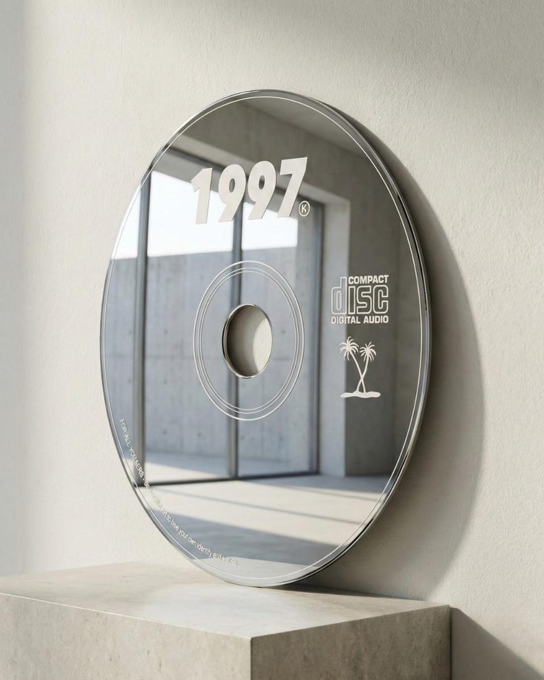 May include: A reflective CD-shaped wall mirror with the year "1997" in white. The mirror has a central hole and circular design elements, with "COMPACT DISC DIGITAL AUDIO" and palm trees. The mirror is displayed on a concrete block.