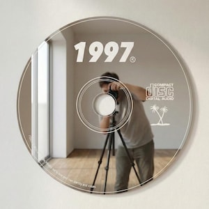 May include: A reflective CD-shaped mirror with the year "1997" printed on it. The mirror reflects a person with a camera on a tripod. The words "COMPACT DISC DIGITAL AUDIO" and two palm trees are also visible.