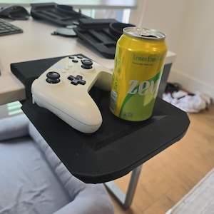 May include: A black desk side tray with a white video game controller and a yellow can of Zevia Lemon Lime soda. The can has the text "Zevia" and "Lemon Lime Soda" printed on it.