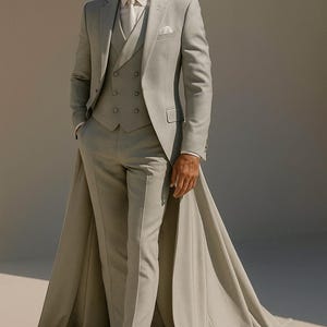 May include: A light grey three-piece suit with a long cape. The suit comprises a blazer, waistcoat, and trousers. The model wears a white shirt, tie, and white boots. The suit is made of a light-coloured fabric. The cape extends to the floor.