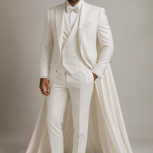 May include: An ivory formal suit comprising a long coat, waistcoat, and trousers. The suit includes a double-breasted waistcoat and a matching bow tie. The long coat has a cape-like extension. The ensemble is finished with white dress shoes.