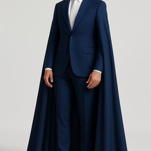 May include: A navy blue suit with a matching cape. The suit includes a blazer, trousers, and a white dress shirt with a tie. The model is wearing white boots. The cape extends to the ankles.