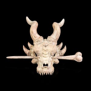 May include: A detailed, off-white skull sculpture with prominent curved horns and sharp teeth. A bone is placed horizontally through the skull. The sculpture is set against a black background, highlighting its intricate details.