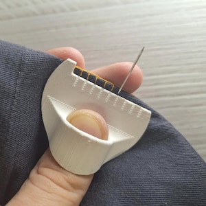 May include: A white 3D-printed sewing tool with a curved edge and evenly spaced notches, designed to fit over a finger. The tool is positioned against a dark gray fabric with yellow stitching, and a thin silver sewing needle is inserted into the fabric.