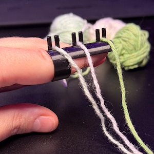 Puede incluir: Un anillo de tensión para hilo de plástico negro con cuatro clavijas verticales en el borde superior. Tres hebras de hilo en verde claro, blanco y morado pálido están enhebradas a través de las clavijas, extendiéndose hacia abajo desde el anillo.