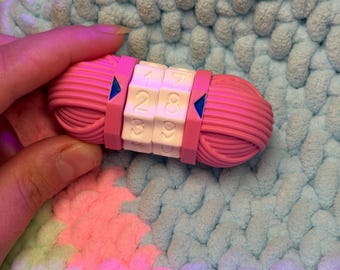 May include: A pink plastic row counter shaped like a yarn ball with two white rotating number dials in the center. The dials feature engraved numbers and are flanked by dark pink bands with blue triangular indicators pointing toward the numbers.