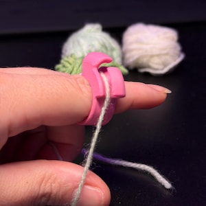 Op de afbeelding: Een felroze plastic garenspanningsring die om een vinger wordt gedragen, voorzien van een klein haakje om een enkele draad lichtgroen garen te geleiden. De ring is ontworpen om een constante spanning te behouden tijdens het breien.