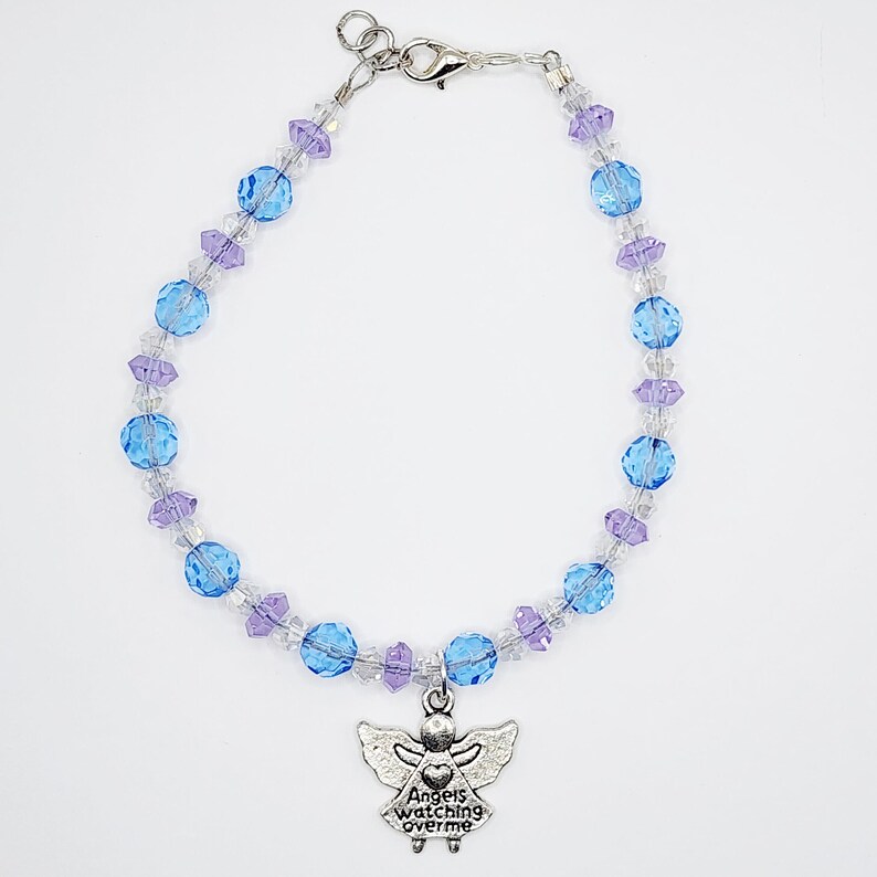 May include: A beaded bracelet featuring blue, purple, and clear beads. A silver-colored angel charm with the words "Angels watching over me" hangs from the bracelet. The bracelet has a silver-colored clasp.