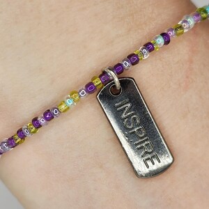 May include: A beaded bracelet with a silver tag that reads "INSPIRE". The bracelet features small, colorful beads in shades of purple, yellow, and clear. The tag is rectangular with rounded corners and hangs from the bracelet.