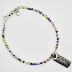 May include: A beaded bracelet with a silver clasp and a rectangular charm. The bracelet features small, colorful beads in shades of purple, green, and blue. The charm is engraved with the word "INSPIRE".