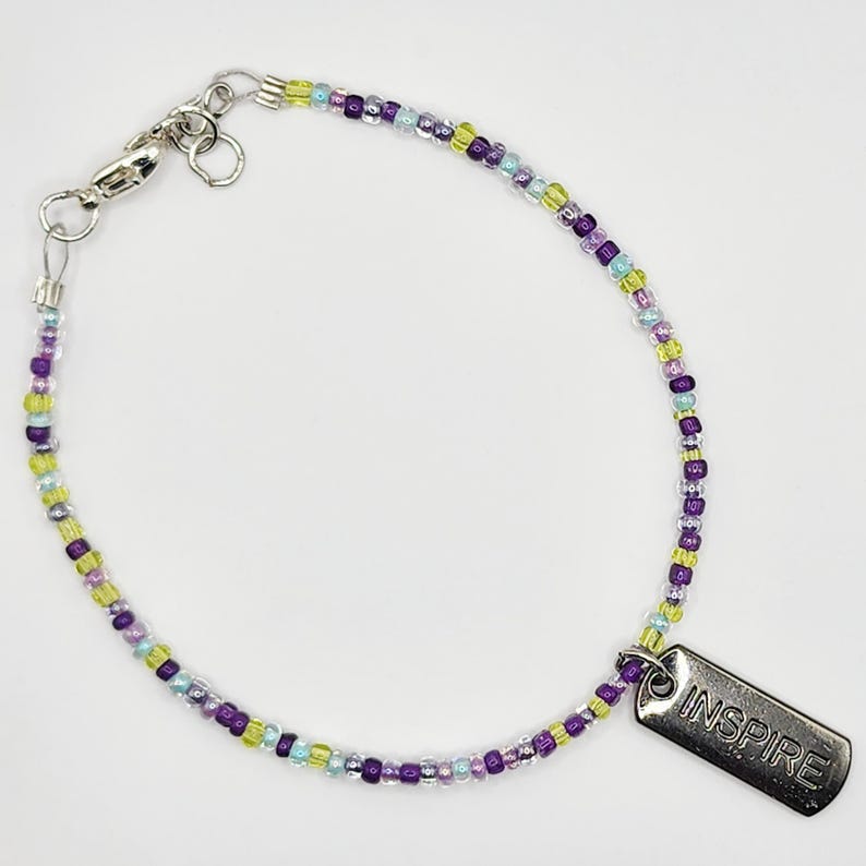 May include: A beaded bracelet with a silver clasp and a rectangular charm. The bracelet features small, colorful beads in shades of purple, green, and blue. The charm is engraved with the word "INSPIRE".