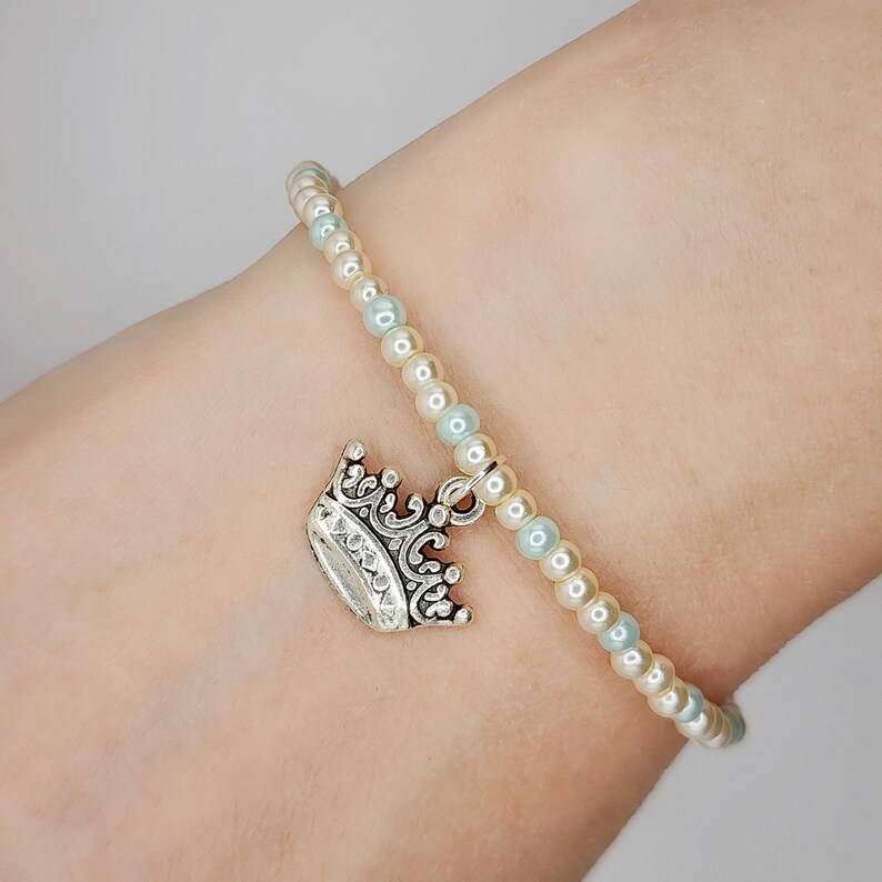 May include: A beaded bracelet with a silver crown charm. The bracelet features alternating cream and light blue beads. The crown charm is detailed with intricate designs. A delicate piece of jewelry.