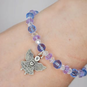 May include: A beaded bracelet with blue, purple, and clear beads. A silver-colored charm in the shape of an angel is attached, with the words "Angels watching over me" inscribed. The bracelet is designed to be worn on the wrist.