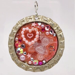 May include: A pink and silver bottle cap pendant with a heart, stars, and rhinestones. The pendant is made of resin and has a shiny finish.