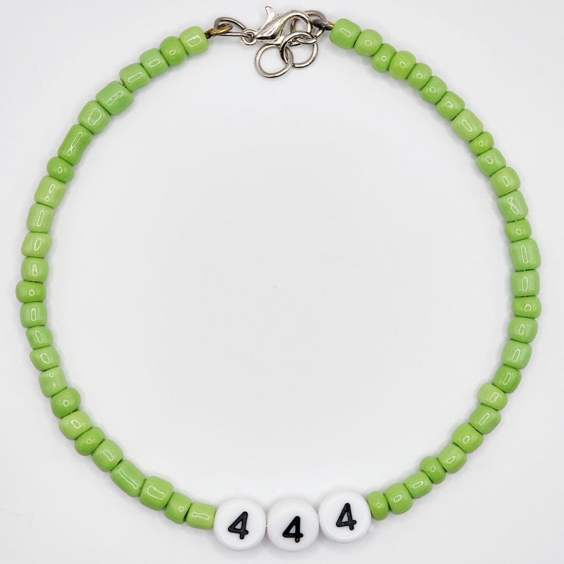 May include: A beaded necklace with light green beads and white letter beads. The letter beads spell out "444" in black letters. The necklace has a silver-colored clasp.