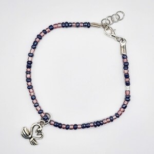 May include: A beaded bracelet featuring alternating dark blue and pink beads. The bracelet has a silver-toned clasp and a charm with two swans. The bracelet is designed to be worn around the wrist.