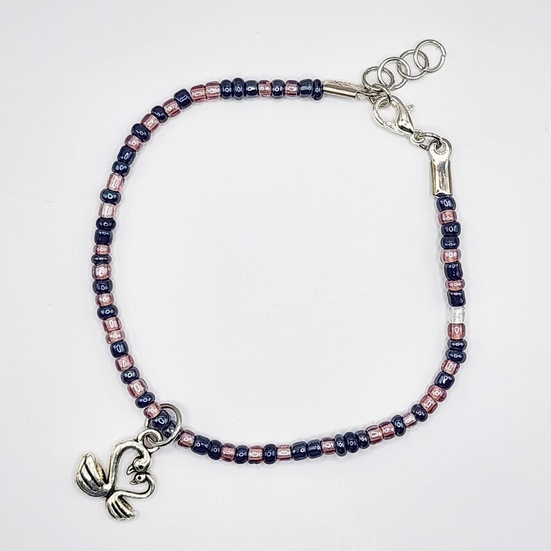 May include: A beaded bracelet featuring alternating dark blue and pink beads. The bracelet has a silver-toned clasp and a charm with two swans. The bracelet is designed to be worn around the wrist.