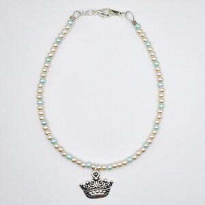 May include: An anklet featuring alternating light blue and cream-colored beads. A silver-colored crown charm hangs from the center. The anklet has a silver-colored clasp.