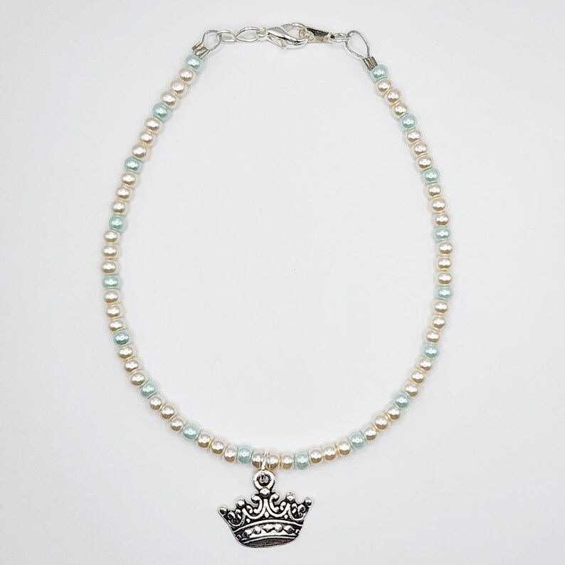 May include: An anklet featuring alternating light blue and cream-colored beads. A silver-colored crown charm hangs from the center. The anklet has a silver-colored clasp.