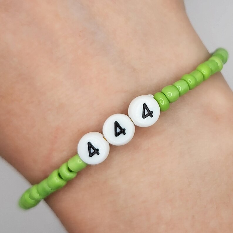 May include: A beaded bracelet with a green base and white beads with the number "4" in black. The bracelet is on a wrist.