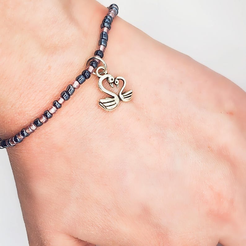 May include: A beaded bracelet with a silver swan charm. The bracelet features a mix of dark blue, pink, and black beads. The swan charm is a detailed, two-swan design. The bracelet is worn on a wrist.