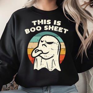 May include: Black sweatshirt with the text "THIS IS BOO SHEET" in white, arched above a cartoon ghost. The ghost is white with a grumpy expression, set against a retro-style circle with horizontal stripes of orange, yellow, blue, and green.