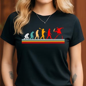 May include: Black t-shirt featuring a retro-style evolution graphic. The graphic shows a progression of figures, from ape-like to a person playing tennis, above horizontal stripes in yellow, orange, red, and green.
