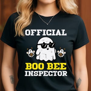 Könnte beinhalten: Schwarzes T-Shirt mit dem Text "OFFICIAL BOO BEE INSPECTOR". Das Design zeigt einen Cartoon-Geist mit Sonnenbrille, flankiert von zwei Cartoon-Bienen. Der Text und die Grafiken sind in Weiß und Gelb.
