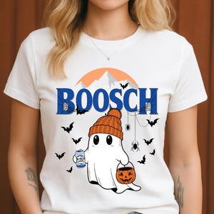 Op de afbeelding: Wit T-shirt met een Halloween-thema. Het ontwerp toont een cartoon spook met een oranje gebreide muts, een bierblikje en een pompoenemmer. Het woord "BOOSCH" staat in blauw gedrukt.