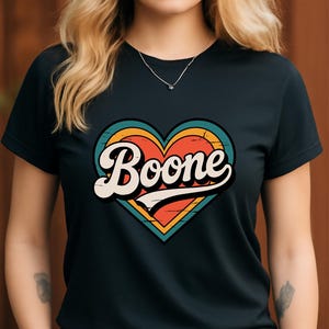 Könnte beinhalten: Schwarzes T-Shirt mit einem Retro-Herz-Design. Das Herz hat Schichten aus Orange, Türkis und Gelb, mit dem Namen "Boone" in weißer Schrift. Ein bequemes und stilvolles T-Shirt.