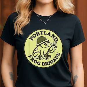 Puede incluir: Camiseta negra con un diseño circular que dice "PORTLAND FROG BRIGADE". El diseño incluye una rana de dibujos animados con casco haciendo el signo de la paz. La rana y el texto son en tonos verdes y amarillos.