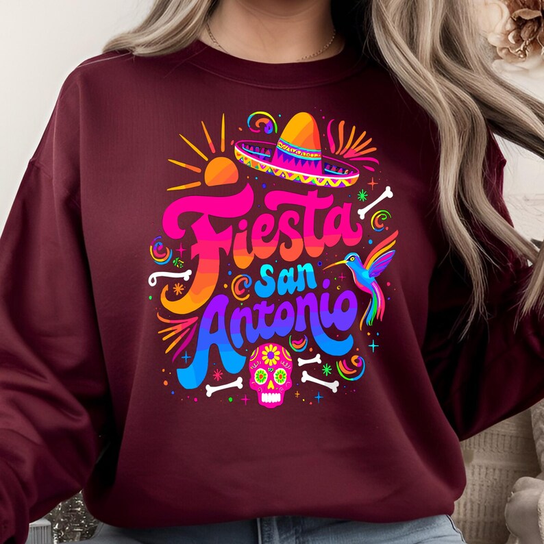 May include: A maroon sweatshirt with a colorful graphic design that says "Fiesta San Antonio". The design includes a sombrero, a hummingbird, and a sugar skull.
