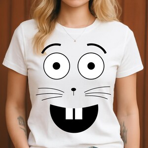 Peut inclure: T-shirt blanc avec un visage de lapin de dessin animé imprimé. Le motif comprend de grands yeux noirs, des sourcils arqués, des moustaches, un nez et une grande bouche ouverte avec deux dents. Le t-shirt a un col rond et des manches courtes.