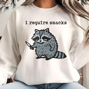May include: A white sweatshirt with the text "i require snacks" above a cartoon raccoon holding a knife. The raccoon is gray and white with a black mask and a striped tail. The sweatshirt is a neutral color.