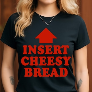 May include: Black t-shirt with a red arrow pointing upwards and the words "INSERT CHEESY BREAD" in red, bold, retro-style font. The shirt has a crew neck and short sleeves.